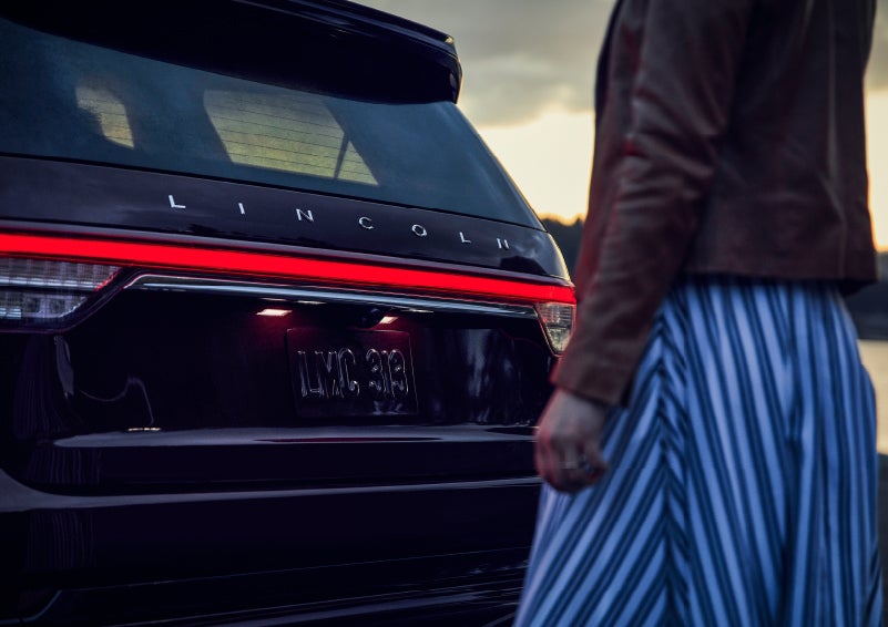 A person is shown near the rear of a 2024 Lincoln Aviator® SUV as the Lincoln Embrace illuminates the rear lights | Mark Ficken Lincoln in Charlotte NC